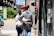 A couple walks down a city street during the day, with their arms around each other. They are surrounded by urban scenery, including greenery, pedestrians, and storefronts. The atmosphere is lively with elements like red awnings, city signs, and a variety of people in the background.