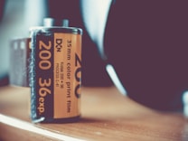 A close-up view of a 35mm Kodak color print film canister with an orange label. The text on the label indicates it is a 200 ISO, 36 exposure film. The canister is placed on a wooden surface, and the background is softly blurred, emphasizing the film roll.