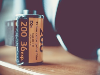 A close-up view of a 35mm Kodak color print film canister with an orange label. The text on the label indicates it is a 200 ISO, 36 exposure film. The canister is placed on a wooden surface, and the background is softly blurred, emphasizing the film roll.