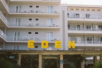 A multi-story hotel building with several balconies, all featuring outdoor chairs and tables. Large yellow letters spelling 'EDEN' are prominently displayed above the entrance. The building is painted in a light color, and the surroundings include some greenery.