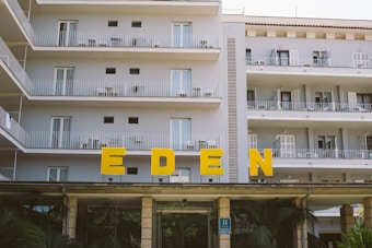 A multi-story hotel building with several balconies, all featuring outdoor chairs and tables. Large yellow letters spelling 'EDEN' are prominently displayed above the entrance. The building is painted in a light color, and the surroundings include some greenery.