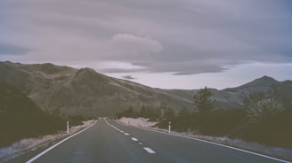 An open road stretching into the horizon, framed by rugged terrain and a moody, overcast sky.