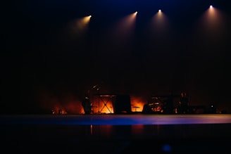 A dark, atmospheric stage with several musical instruments illuminated by soft spotlights. The scene is minimally lit with warm, ambient lighting creating a moody and contemplative setting.