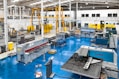 A spacious industrial workshop with various pieces of machinery and equipment organized throughout the area. The floor is a bright blue, and several large machines are positioned around the space, including a metal press and a Euromac machine. Overhead, there are support beams and lighting fixtures, and a yellow overhead crane system is visible. The walls are white with large windows, and there are several workbenches around the area. A few people are working in the background.