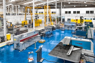 A spacious industrial workshop with various pieces of machinery and equipment organized throughout the area. The floor is a bright blue, and several large machines are positioned around the space, including a metal press and a Euromac machine. Overhead, there are support beams and lighting fixtures, and a yellow overhead crane system is visible. The walls are white with large windows, and there are several workbenches around the area. A few people are working in the background.