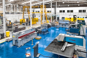 A spacious industrial workshop with various pieces of machinery and equipment organized throughout the area. The floor is a bright blue, and several large machines are positioned around the space, including a metal press and a Euromac machine. Overhead, there are support beams and lighting fixtures, and a yellow overhead crane system is visible. The walls are white with large windows, and there are several workbenches around the area. A few people are working in the background.