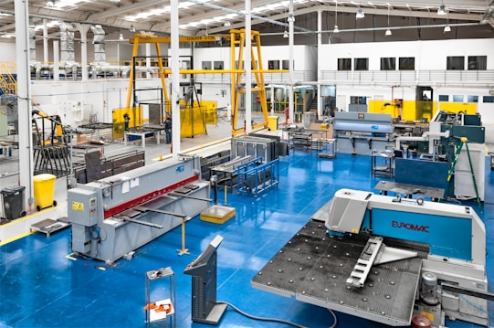 A spacious industrial workshop with various pieces of machinery and equipment organized throughout the area. The floor is a bright blue, and several large machines are positioned around the space, including a metal press and a Euromac machine. Overhead, there are support beams and lighting fixtures, and a yellow overhead crane system is visible. The walls are white with large windows, and there are several workbenches around the area. A few people are working in the background.