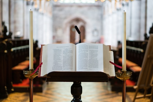 An open book is placed on a lectern in the center of a church aisle. Two tall candles flank the book, and a microphone is visible above. The background reveals the ornate interior architecture of the church, with rows of pews and a blurred, richly decorated wall.