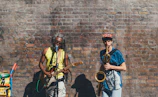 Two musicians perform against a weathered brick wall. One plays a guitar and sings into a microphone, wearing a bright yellow tank top. The other plays a saxophone, wearing a colorful cap and blue shirt. A sound system with colorful stickers is positioned nearby.
