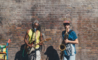 Two musicians perform against a weathered brick wall. One plays a guitar and sings into a microphone, wearing a bright yellow tank top. The other plays a saxophone, wearing a colorful cap and blue shirt. A sound system with colorful stickers is positioned nearby.