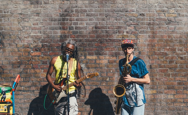 Two musicians perform against a weathered brick wall. One plays a guitar and sings into a microphone, wearing a bright yellow tank top. The other plays a saxophone, wearing a colorful cap and blue shirt. A sound system with colorful stickers is positioned nearby.