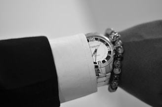 A close-up view of a wrist wearing a silver analog wristwatch with a white face and bold black numbers. The wrist is adorned with a formal long sleeve black and white shirt and a beaded bracelet.