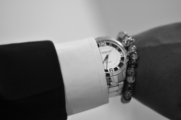 A close-up view of a wrist wearing a silver analog wristwatch with a white face and bold black numbers. The wrist is adorned with a formal long sleeve black and white shirt and a beaded bracelet.
