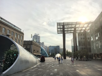 A modern urban scene with a sunlit plaza featuring contemporary architecture and sculptures. Buildings in the background include recognizable brand signage. Several people are walking around the area.