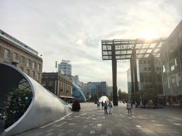 A modern urban scene with a sunlit plaza featuring contemporary architecture and sculptures. Buildings in the background include recognizable brand signage. Several people are walking around the area.