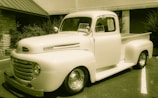 A vintage pickup truck is parked outside a building, with signs in the windows indicating it is for sale. The truck has a classic rounded design, prominent front grille, and is in a cream color. It is situated on an asphalt surface with some greenery in the background.