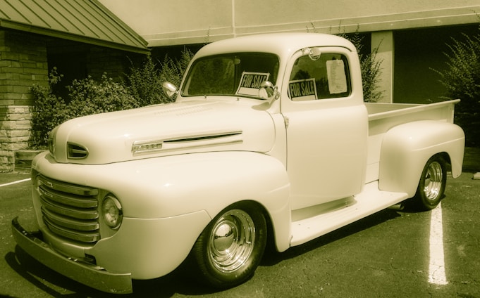 A vintage pickup truck is parked outside a building, with signs in the windows indicating it is for sale. The truck has a classic rounded design, prominent front grille, and is in a cream color. It is situated on an asphalt surface with some greenery in the background.