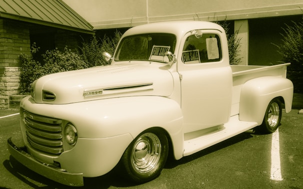 A vintage pickup truck is parked outside a building, with signs in the windows indicating it is for sale. The truck has a classic rounded design, prominent front grille, and is in a cream color. It is situated on an asphalt surface with some greenery in the background.