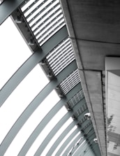 A perspective view of an architectural structure showcasing a series of curved steel beams forming an arc. The beams are complemented by slatted panels and lights attached along the interior. The image features a modern, industrial design with a focus on geometric lines and patterns.