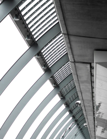 A perspective view of an architectural structure showcasing a series of curved steel beams forming an arc. The beams are complemented by slatted panels and lights attached along the interior. The image features a modern, industrial design with a focus on geometric lines and patterns.