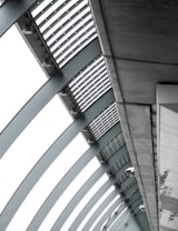 A perspective view of an architectural structure showcasing a series of curved steel beams forming an arc. The beams are complemented by slatted panels and lights attached along the interior. The image features a modern, industrial design with a focus on geometric lines and patterns.