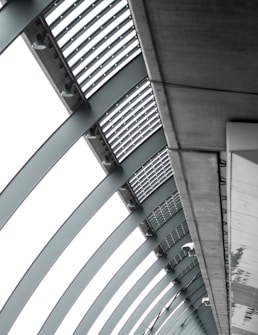 A perspective view of an architectural structure showcasing a series of curved steel beams forming an arc. The beams are complemented by slatted panels and lights attached along the interior. The image features a modern, industrial design with a focus on geometric lines and patterns.