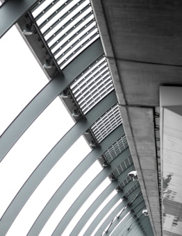 A perspective view of an architectural structure showcasing a series of curved steel beams forming an arc. The beams are complemented by slatted panels and lights attached along the interior. The image features a modern, industrial design with a focus on geometric lines and patterns.