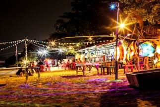 A vibrant outdoor scene at night with colorful lights and festive decorations. Several people are sitting on red plastic chairs around tables set on sandy ground. Strings of flag bunting and brightly illuminated structures create a lively and inviting atmosphere, while trees and a small building are visible in the background.