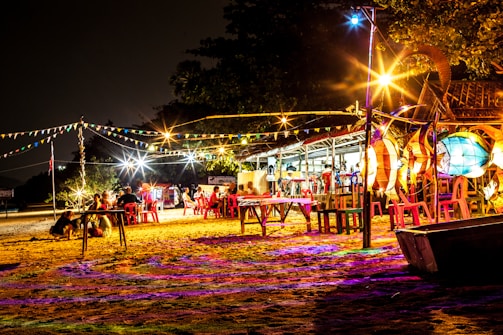 A vibrant outdoor scene at night with colorful lights and festive decorations. Several people are sitting on red plastic chairs around tables set on sandy ground. Strings of flag bunting and brightly illuminated structures create a lively and inviting atmosphere, while trees and a small building are visible in the background.