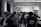 A black and white image capturing a social gathering or networking event in an indoor setting. Many people are mingling, standing, and conversing with one another. The environment features large structural columns, fluorescent ceiling lights, and industrial-style architecture. Attendees are casually dressed and some individuals appear to be holding drinks or plates.
