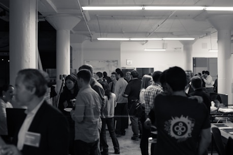 A black and white image capturing a social gathering or networking event in an indoor setting. Many people are mingling, standing, and conversing with one another. The environment features large structural columns, fluorescent ceiling lights, and industrial-style architecture. Attendees are casually dressed and some individuals appear to be holding drinks or plates.