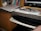 A cluttered desk area featuring a stack of books on a shelf, papers and a binder on the countertop, with a pair of glasses resting on top. There's a keyboard on a slide-out tray underneath the counter and part of a computer monitor is visible on the side.