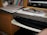 A cluttered desk area featuring a stack of books on a shelf, papers and a binder on the countertop, with a pair of glasses resting on top. There's a keyboard on a slide-out tray underneath the counter and part of a computer monitor is visible on the side.