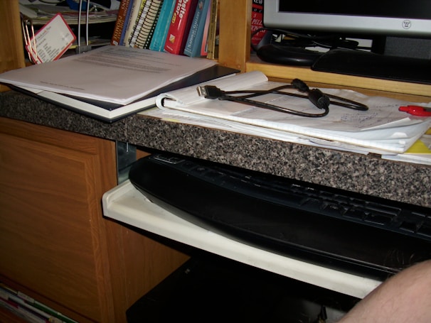 A cluttered desk area featuring a stack of books on a shelf, papers and a binder on the countertop, with a pair of glasses resting on top. There's a keyboard on a slide-out tray underneath the counter and part of a computer monitor is visible on the side.
