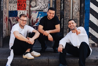 Three men are sitting on a sidewalk in front of a wall covered with various graffiti and posters. They appear relaxed and are smiling, conveying a sense of camaraderie. The men are dressed casually, with two wearing white shirts and the one in the middle wearing a black shirt.