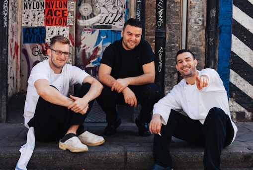 Three men are sitting on a sidewalk in front of a wall covered with various graffiti and posters. They appear relaxed and are smiling, conveying a sense of camaraderie. The men are dressed casually, with two wearing white shirts and the one in the middle wearing a black shirt.