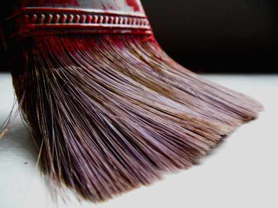 Close-up view of the bristles of a paintbrush. The bristles have a reddish tint towards the top near the metal ferrule and gradually shift to a muted brown color towards the tip. The intricate details of individual bristles are visible, highlighting their texture and alignment. The metal ferrule is engraved with a pattern, and the overall lighting suggests a soft, natural ambiance.