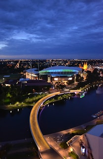 An evening cityscape featuring a stadium illuminated with colorful lights, surrounded by trees and buildings. A curved, well-lit pedestrian bridge crosses over a river. The sky is darkening with a deep blue hue, contrasting against city lights.