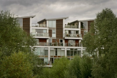 A modern apartment building surrounded by lush green trees under a cloudy sky. The structure features multiple floors with large glass windows and wooden paneling. Balconies are present on each floor, some adorned with potted plants.