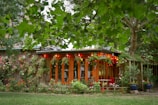 A cozy wooden garden house adorned with colorful flowering plants and string lights, surrounded by lush green trees and bushes. The setting creates a serene and welcoming outdoor space, with garden furniture indicating a place to relax and enjoy nature.