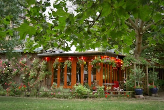 A cozy wooden garden house adorned with colorful flowering plants and string lights, surrounded by lush green trees and bushes. The setting creates a serene and welcoming outdoor space, with garden furniture indicating a place to relax and enjoy nature.
