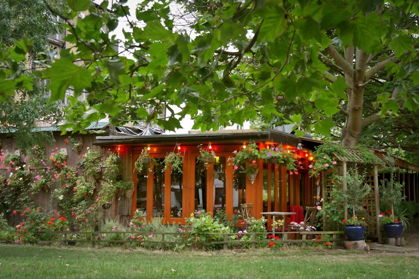 A cozy wooden garden house adorned with colorful flowering plants and string lights, surrounded by lush green trees and bushes. The setting creates a serene and welcoming outdoor space, with garden furniture indicating a place to relax and enjoy nature.