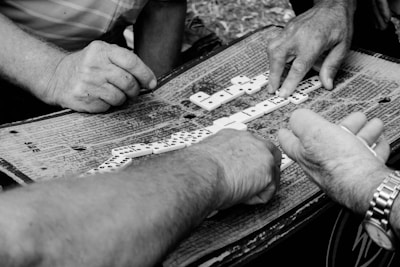 Several hands are gathered around a table engaged in a game of dominoes. The table surface is worn and covered in markings, indicating frequent use. The scene captures the intensity and focus of the players as they strategically place the pieces.