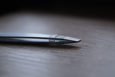 A close-up view of a pair of metallic tweezers with a sleek design lying on a wooden surface. The focus is on the pointed end, showcasing the fine tips and reflective surface.