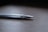 A close-up view of a pair of metallic tweezers with a sleek design lying on a wooden surface. The focus is on the pointed end, showcasing the fine tips and reflective surface.