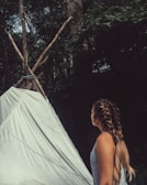 A person with long braided hair, wearing a tank top, is observing a simple teepee made of tied logs and a white sheet. The background is filled with dense green foliage, indicating a forest setting.