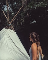 A person with long braided hair, wearing a tank top, is observing a simple teepee made of tied logs and a white sheet. The background is filled with dense green foliage, indicating a forest setting.