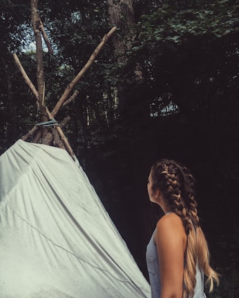 A person with long braided hair, wearing a tank top, is observing a simple teepee made of tied logs and a white sheet. The background is filled with dense green foliage, indicating a forest setting.