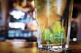 A close-up view of a glass filled with a refreshing beverage, featuring lime wedges submerged in the liquid. The background appears to be a lively and slightly blurred bar or restaurant setting, indicating a vibrant atmosphere.