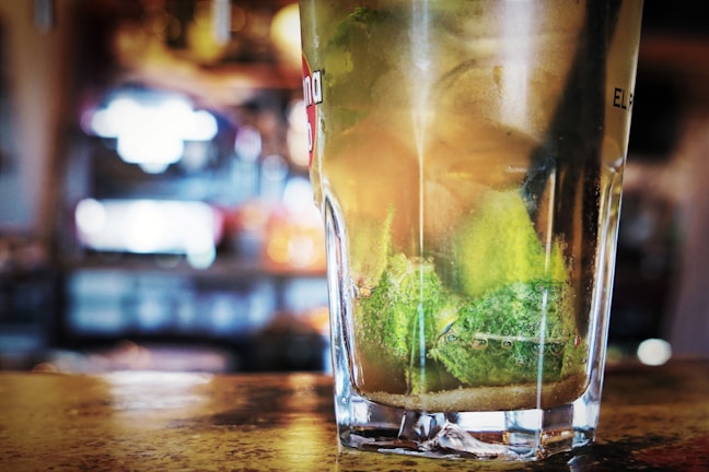 A close-up view of a glass filled with a refreshing beverage, featuring lime wedges submerged in the liquid. The background appears to be a lively and slightly blurred bar or restaurant setting, indicating a vibrant atmosphere.